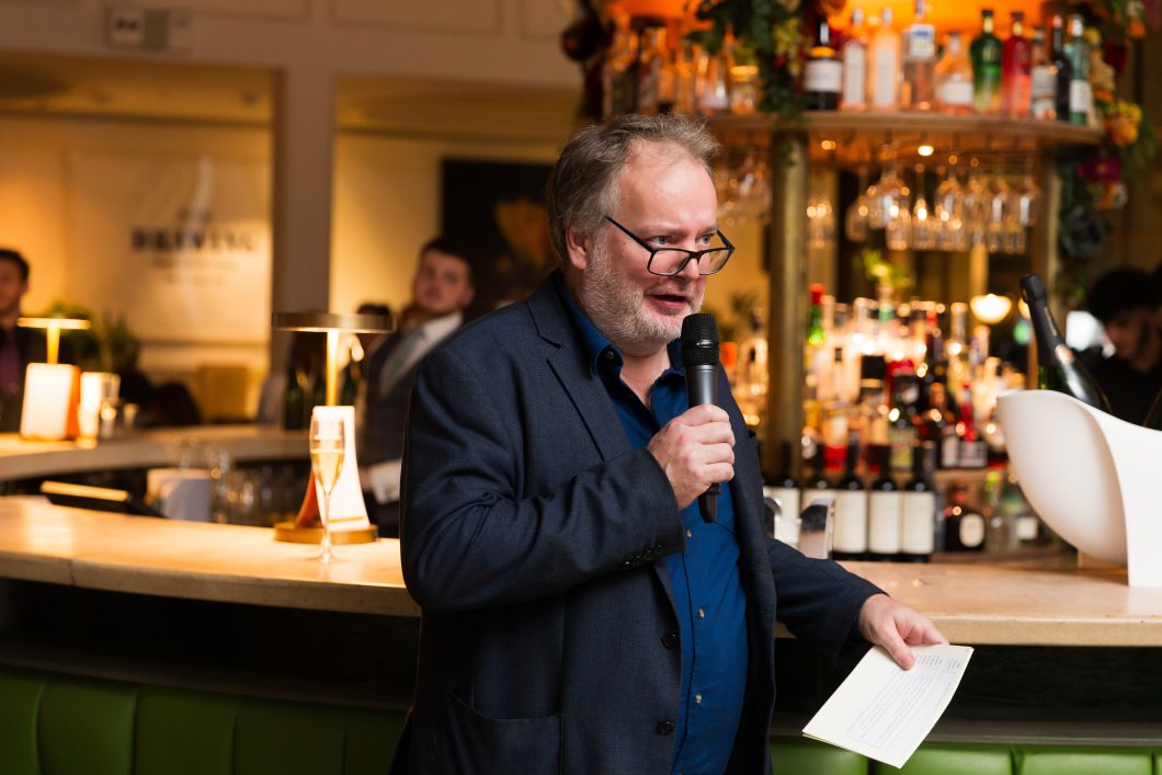 Bill Knott stands in a restaurant, holding a microphone in one hand and cue cards in the other. He is mid speech.