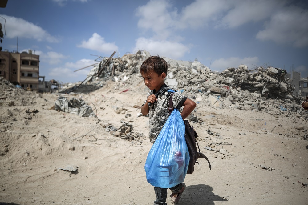 A small child stands in front of buildings destroyed to rubble in Gaza, holding his belongings in a blue carrier bag.