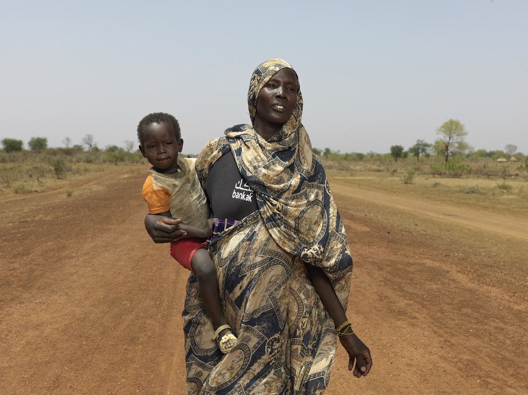 Nyibol, from Sudan, holding her child while walking on sandy road in South Sudan.