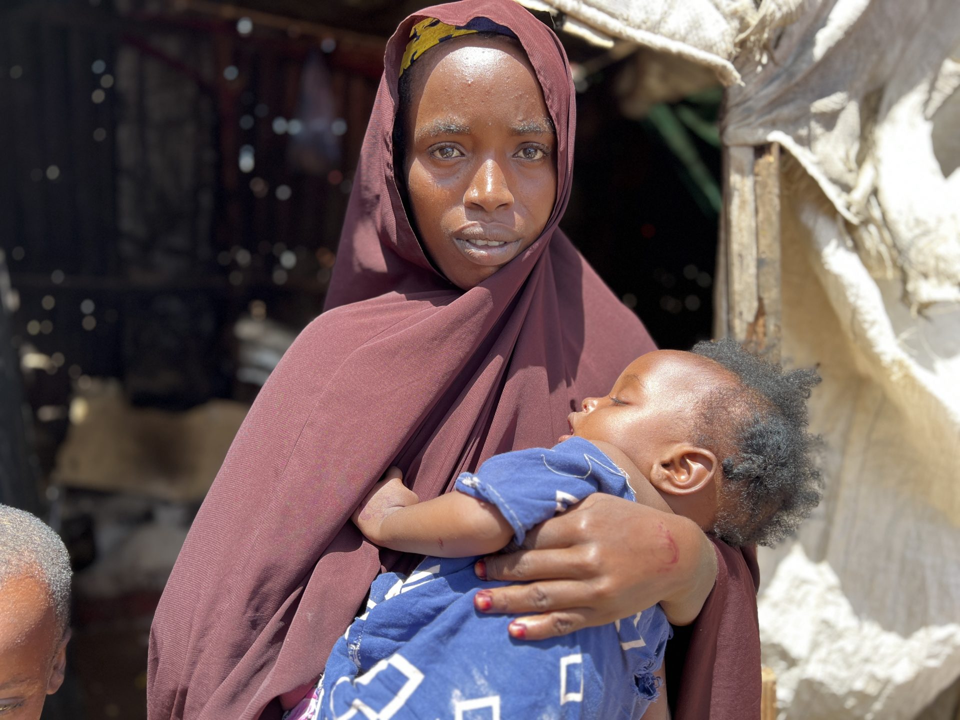 Mother in Somalia in brown headscarf holding her child outside home.