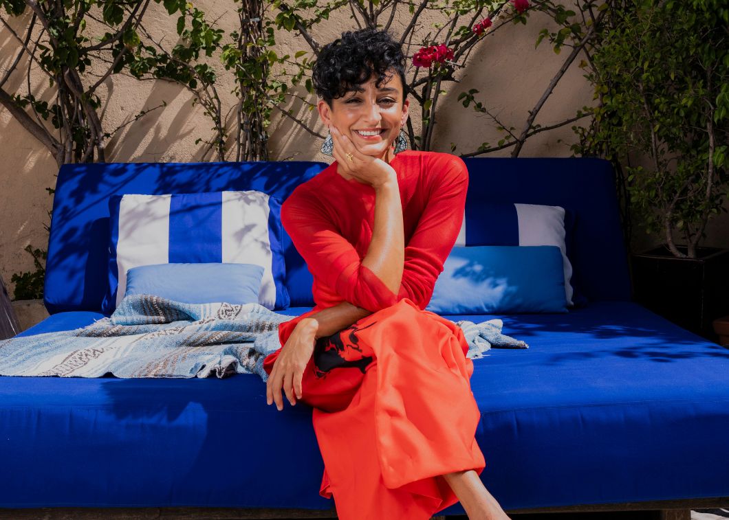 Too Many Critics host Leyla Kazim sits on sofa in bright red dress.