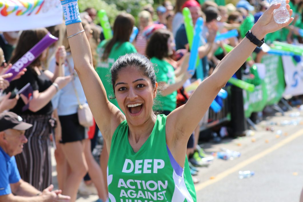 Woman wearing an Action Against Hunger fundraising vest with arms in air smiling while running marathon.